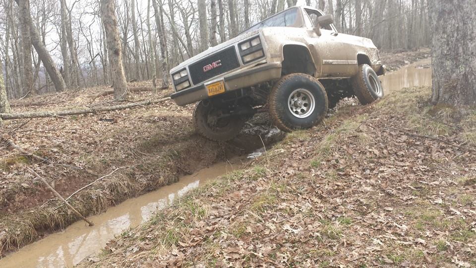 Mud-splattered GMC pickup truck with large tires navigating a muddy ditch on a wooded hill