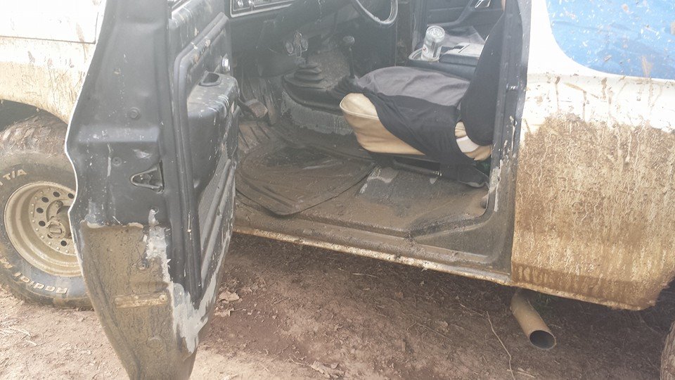 Mud splattered on the door, floor, and exterior of an off-road vehicle. The driver's side door is open, showing the interior with floor mats and seats