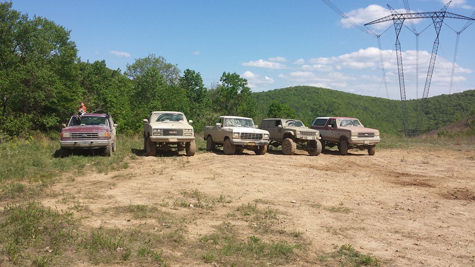 five lifted trucks and SUVs parked on a dirt path with trees and hills in the background