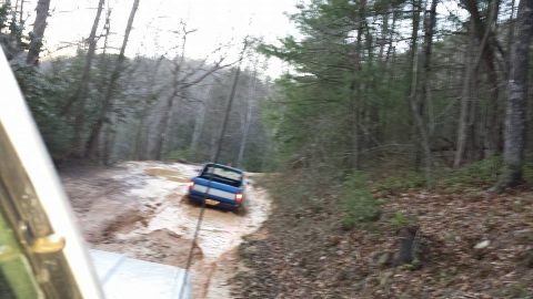 1979 Ford Bronco - A blue pickup truck navigates a muddy trail through a forest, with another vehicle close behind.