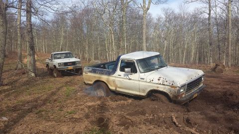 1979 Ford Bronco - Two trucks are stuck in the mud on a wooded trail, with one attempting to pull the other out.