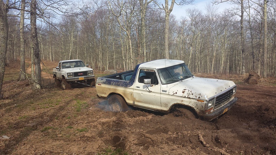 White Ford Bronco stuck in mud, with a white GMC truck pulling it
