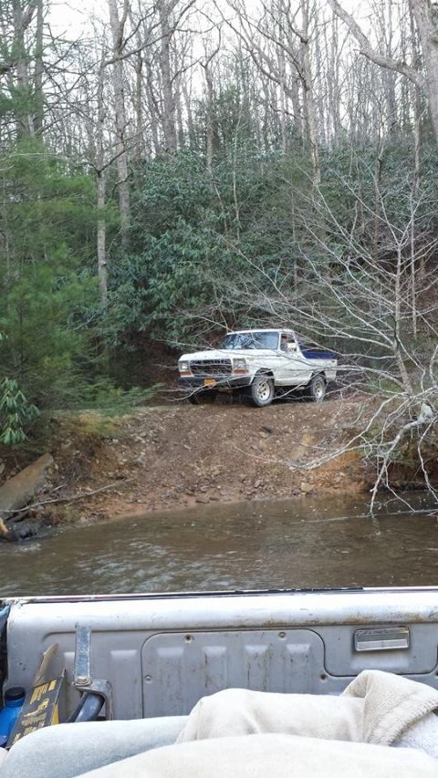 1979 Ford Bronco - A white Ford pickup truck navigates a muddy path beside a stream in a wooded area.