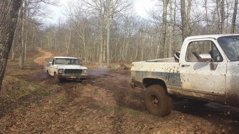 1979 Ford Bronco - Two trucks navigate a muddy trail through a wooded area, kicking up dirt and debris.