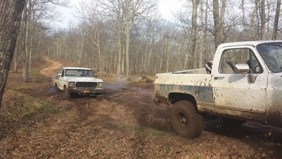 White 1979 Ford pickup truck driving through mud in a forest, with another truck visible