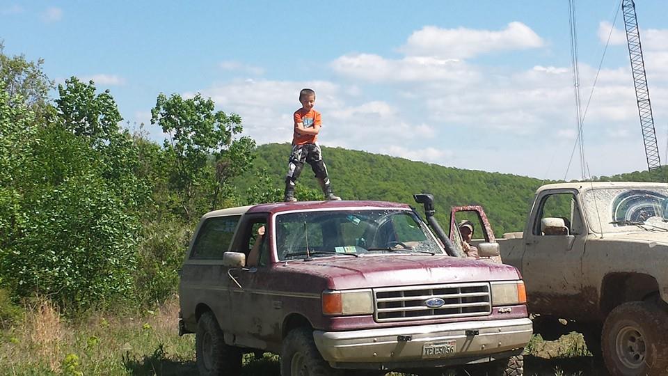 Maroon Ford Bronco with a child standing on its roof, parked next to a white pickup truck