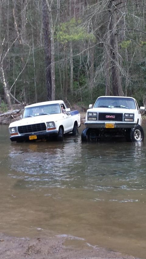 1979 Ford Bronco - Two trucks are partially submerged in a shallow creek bed surrounded by trees.