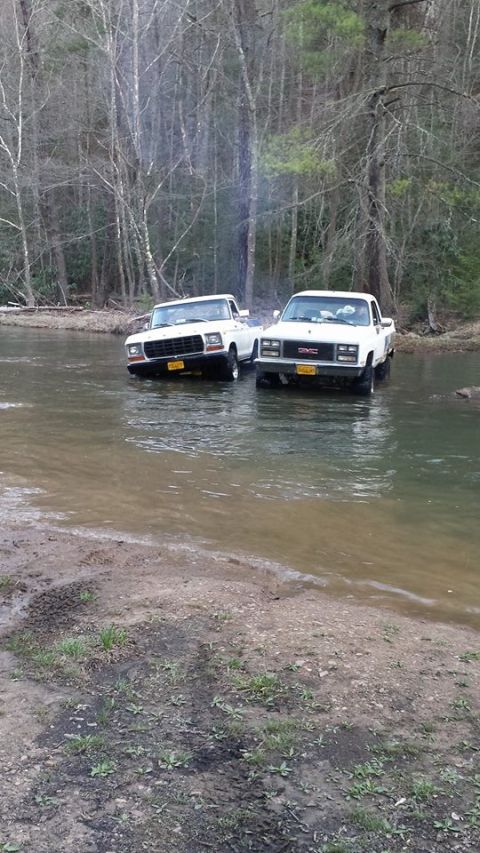 1979 Ford Bronco - Two pickup trucks, a Ford and a GMC, drive through a shallow river surrounded by trees.