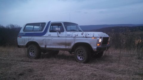 1979 Ford Bronco - A white and blue Ford Bronco sits in a field with hills in the background under a cloudy sky.