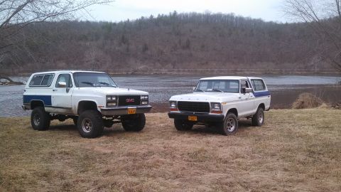 1979 Ford Bronco - Two classic SUVs, a Ford Bronco and a GMC Jimmy, sit parked near a body of water on a cloudy day.