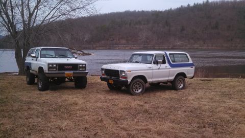 1979 Ford Bronco - Two SUVs, a 1978 Ford Bronco and a GMC, are parked near a lake with wooded hills in the background.