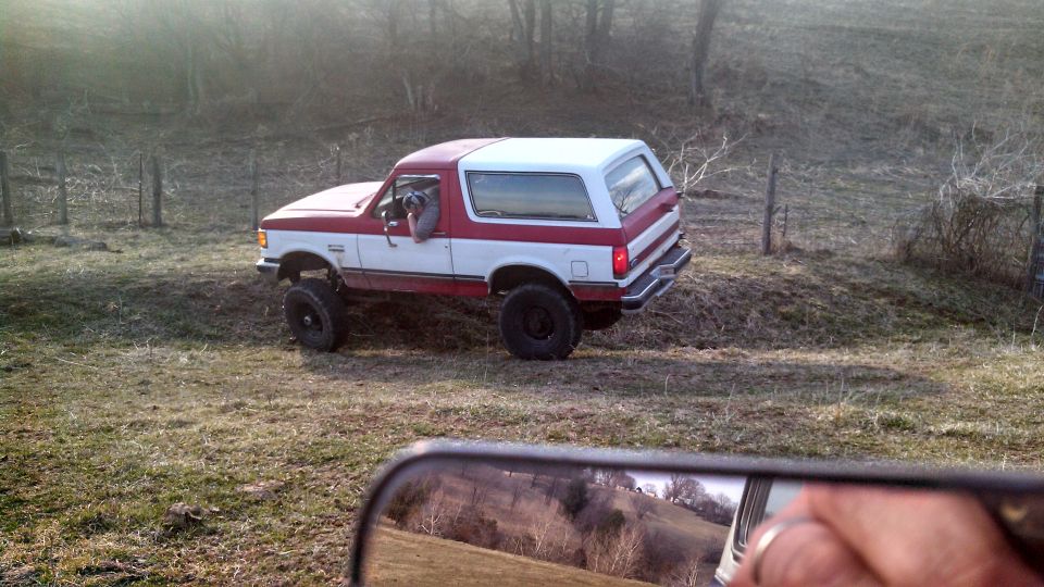 Red and white Ford Bronco with lifted suspension driving off-road. A dog is visible in the driver's seat