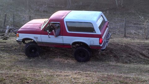 1979 Ford Bronco - This two-tone Ford Bronco is navigating a grassy, uneven slope with trees in the background.