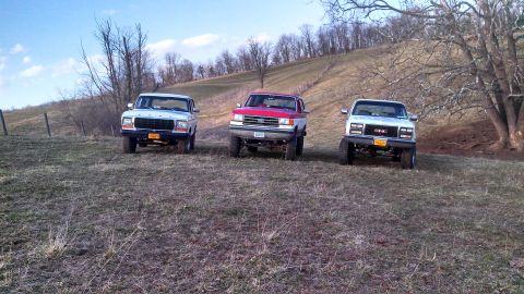 1979 Ford Bronco - Three trucks are parked on a grassy hillside under a partly cloudy sky.