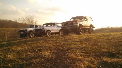 1979 Ford Bronco - Three SUVs are parked on a grassy hill during sunset, with the sun low in the sky.