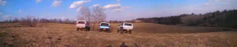 1979 Ford Bronco - Three SUVs are parked in a wide, open field with rolling hills and trees in the background.
