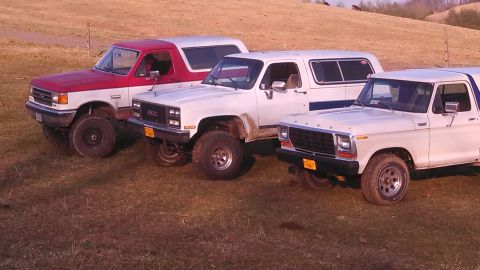 1979 Ford Bronco - Three SUVs, including a 1979 Ford Bronco, are parked side-by-side in a grassy field.