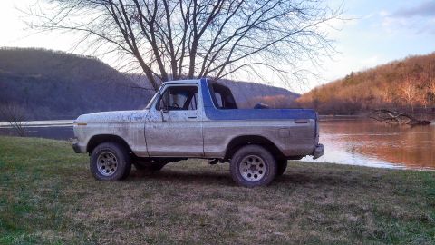 1979 Ford Bronco - A 1979 Ford Bronco sits on grassy terrain next to a calm river under a tree.