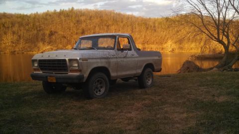 1979 Ford Bronco - A dusty Ford Bronco sits on a grassy bank next to a calm body of water.