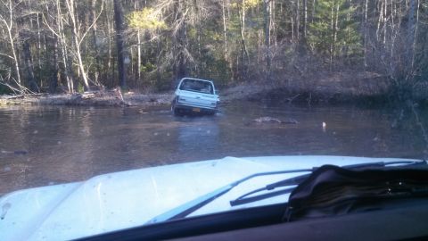 1979 Ford Bronco - Pickup truck navigating through a shallow body of water in a wooded area.