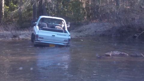 1979 Ford Bronco - A white GMC pickup truck navigates through a muddy, shallow body of water surrounded by trees.