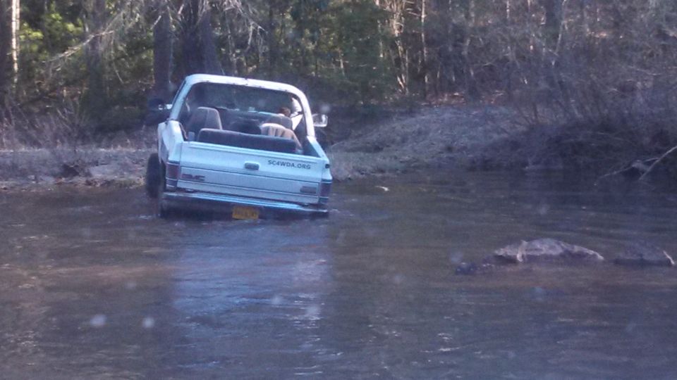 White GMC pickup truck driving through shallow water, trees in background