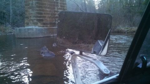 1979 Ford Bronco - The vehicle's interior is visible, with the side mirror reflecting the surroundings as it drives through water.