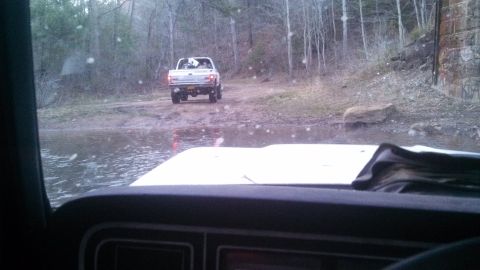 1979 Ford Bronco - A white pickup truck navigates a water-filled dirt road in a wooded area.