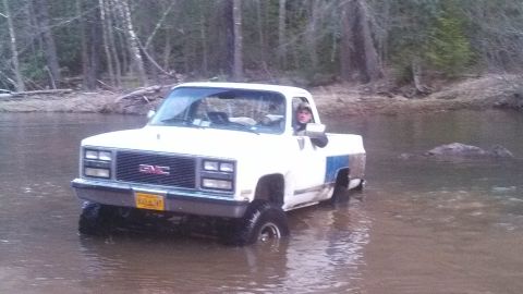 1979 Ford Bronco - GMC pickup truck navigates a river crossing in a wooded area.
