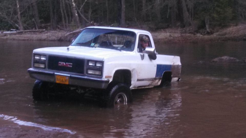 White GMC pickup truck with blue stripe driving in shallow water, driver visible