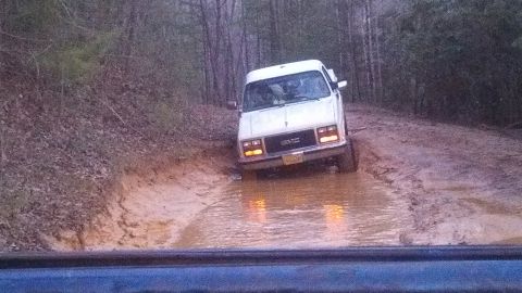 1979 Ford Bronco - A white van navigates a muddy trail, its tires partially submerged in water.