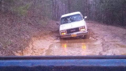 1979 Ford Bronco - A white GMC van struggles through a muddy, water-filled trail surrounded by trees.