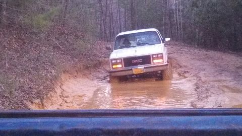 1979 Ford Bronco - White GMC pickup truck navigates a muddy off-road trail.
