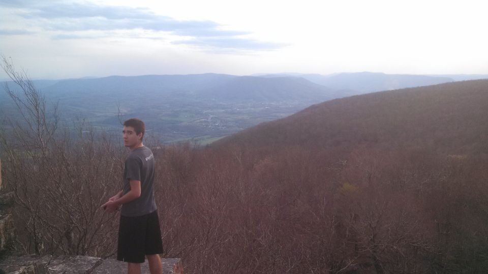 guy looking back over shoulder on rocky ledge with valley and mountains in background