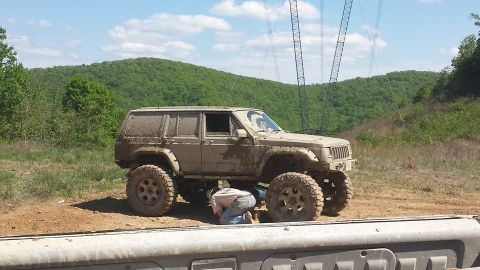 1979 Ford Bronco - A person works on a lifted, mud-covered Jeep Cherokee XJ with large off-road tires, parked on a dirt path with power lines in the backgroun…