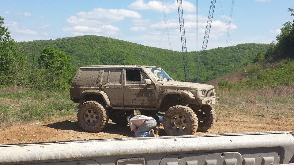 Mud-covered Jeep Cherokee XJ with large tires, being worked on by a person underneath