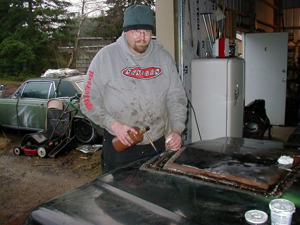 Man with torch and bottle working on rusty cutout in black hood panel at garage, green car behind