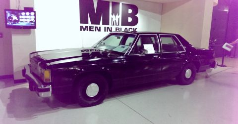 2000 Mercury Grand Marquis - A black sedan with silver stripes on the hood is parked indoors near a 'Men in Black' display.