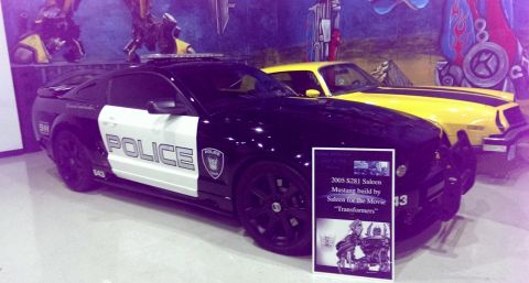 2000 Mercury Grand Marquis - Two cars, a police-liveried Mustang and a yellow car, are displayed indoors with painted murals in the background.