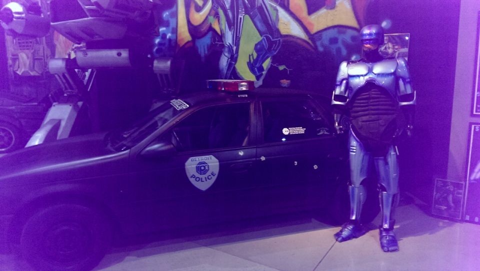 Black police car with a Detroit Police badge, bullet holes, and a red light bar, parked next to a Robocop costume