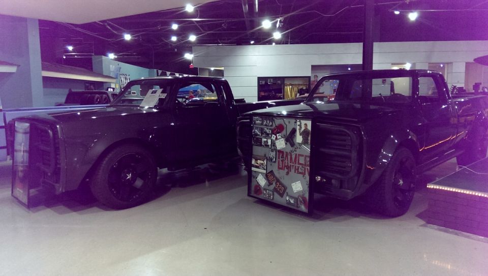 Two black custom pickup trucks side-by-side. One has a display board on its front fender with stickers and text that reads 'BRUCE BAPTIST'