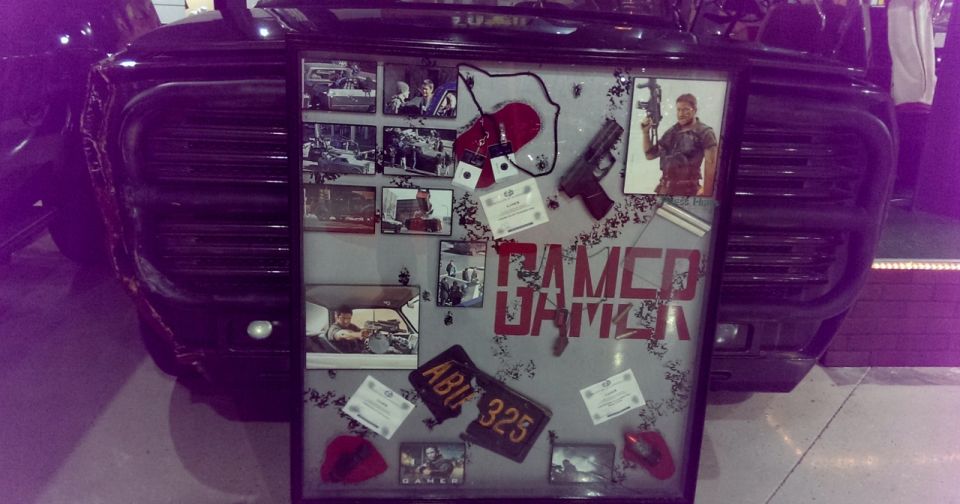 Display board with photos, a gun, and 'GAMER' text, mounted on a dark vehicle's grille