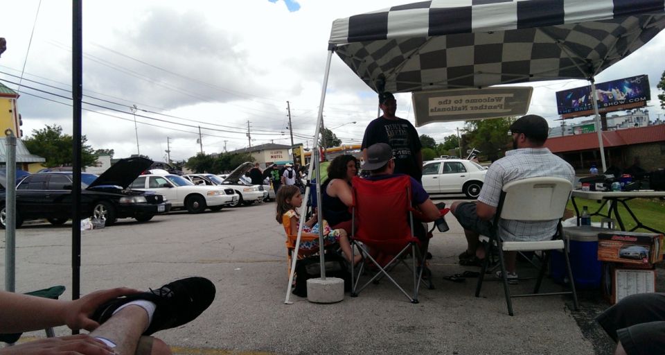People gathered under a checkered tent at a car show, with several cars parked in the background. A 2000 Mercury Grand Marquis is visible