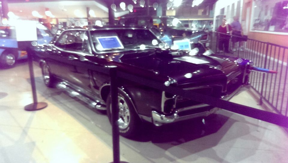 Black classic coupe with chrome trim, side pipes, and hood scoop. Displayed indoors with stanchions