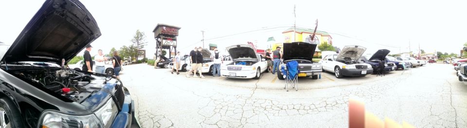 Several cars with hoods open parked in a lot, some people standing around