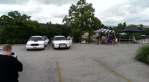 2000 Mercury Grand Marquis - Two police interceptor sedans are parked side-by-side in a lot, with people socializing under tents in the background.