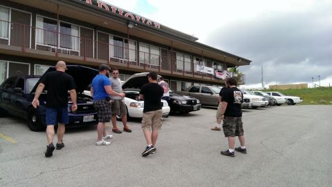 2000 Mercury Grand Marquis - A group of people gather around a police car with its hood open, parked outside a motel with other vehicles in the background.