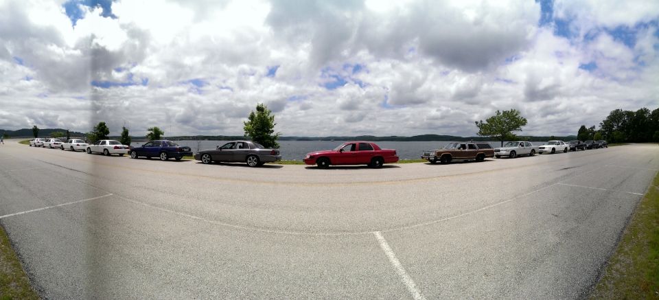 Line of sedans and a station wagon parked on asphalt by a lake under a cloudy sky