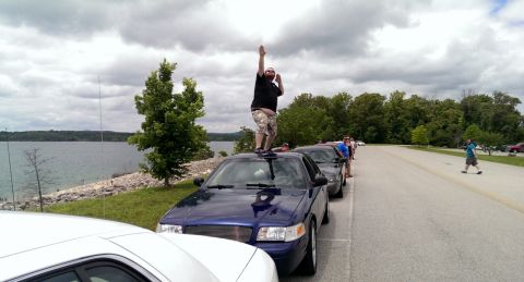 2000 Mercury Grand Marquis - A man stands atop a blue 2000 Mercury Grand Marquis, parked in a line of cars next to a lake.