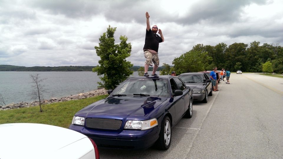 Blue 2000 Mercury Grand Marquis with a man standing on its roof, parked in a line of cars by a lake